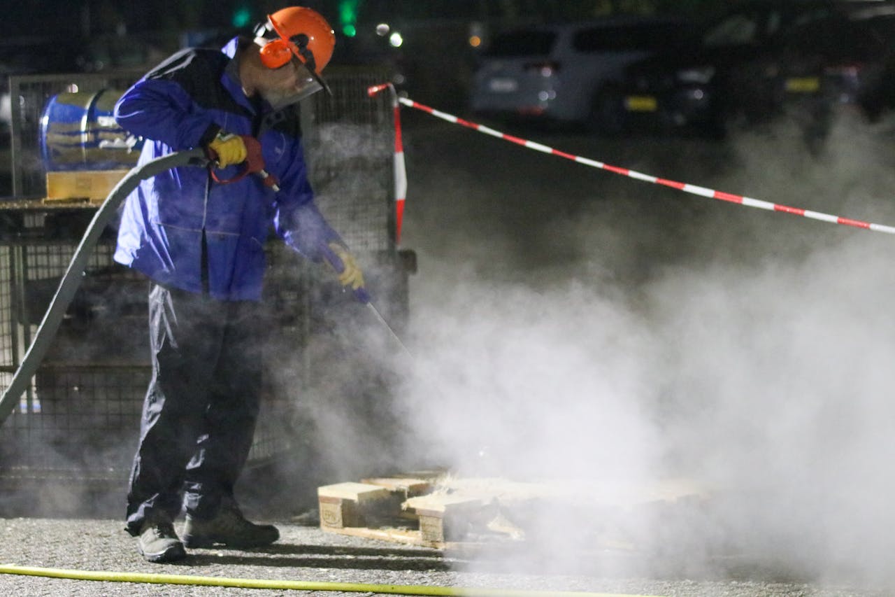 gallery-4 Man in protective gear using a high-pressure washer amid steam at night in an industrial setting.